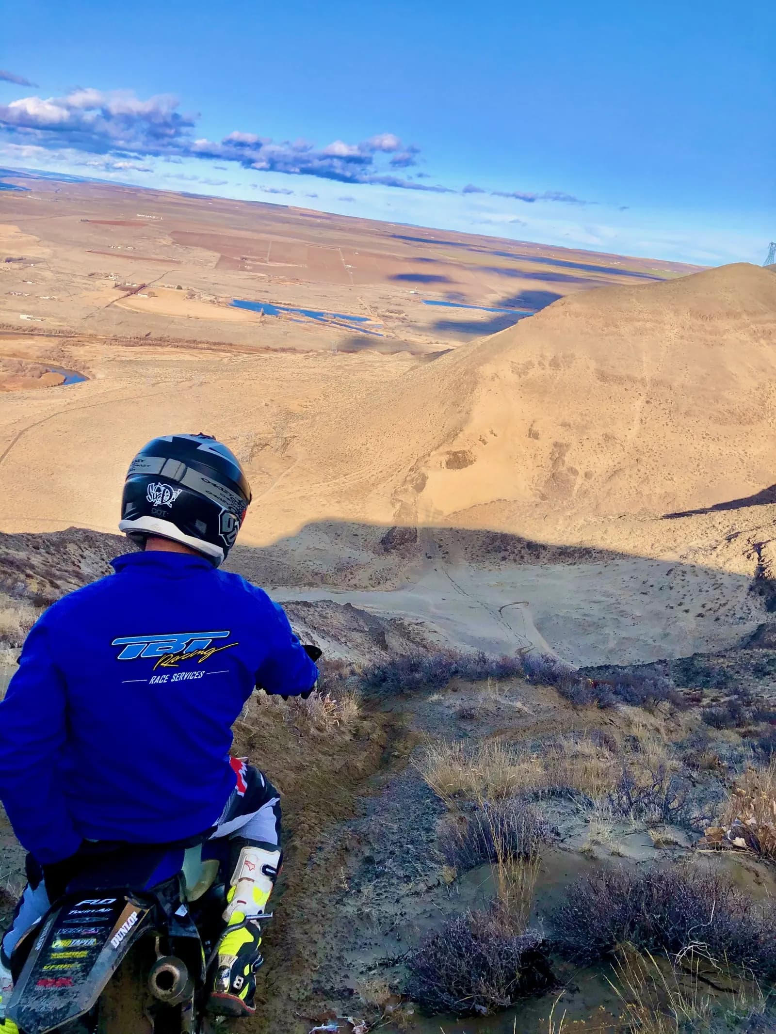A rider in TBT Racing gear pauses on a high ridge, looking out over the valley and the road ahead.